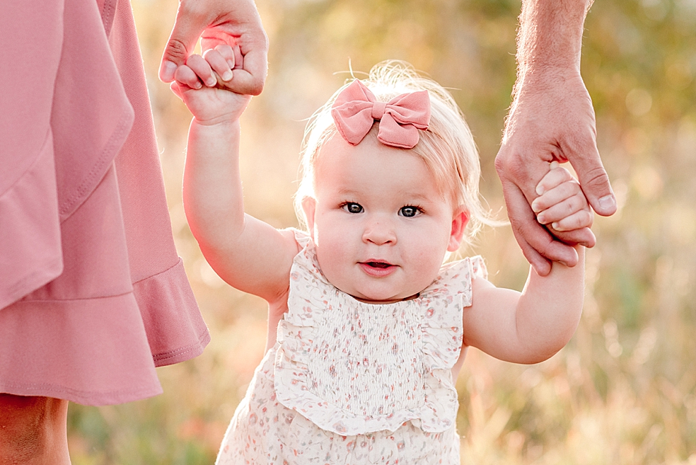 Baby girl in pink bow holding parents hands | Photo by Jessica Lee Photography