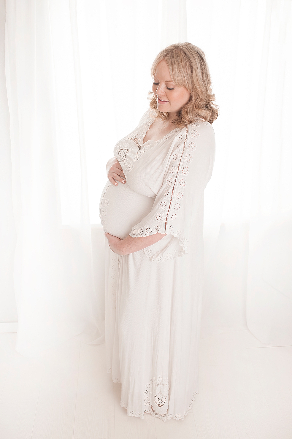 Mom to be in long white dress by a window with sheer drapes | Photo by Jessica Lee Photography
