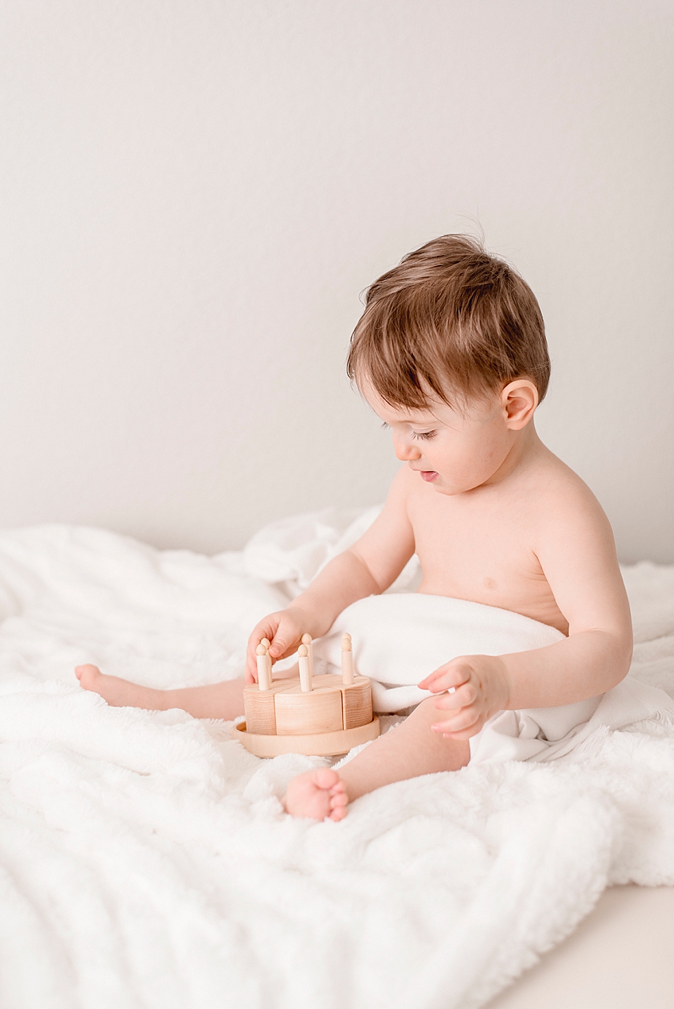 Baby boy on soft white blanket playing with wooden cake toy | Photo by Jessica Lee Photography